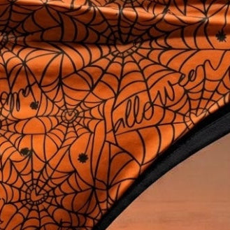 Orange bikini bottoms with a black spiderweb and spider graphic print, displayed on a light wooden surface against a dark gray background.