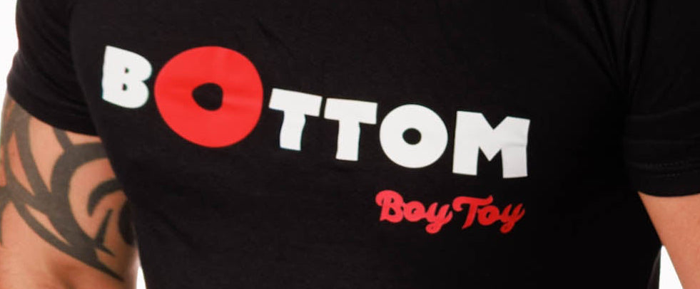 A man wearing a black short-sleeve t-shirt with a graphic on the chest that reads 'BOTTOM BoyToy' in white and red, paired with light-wash distressed jeans against a plain white background.