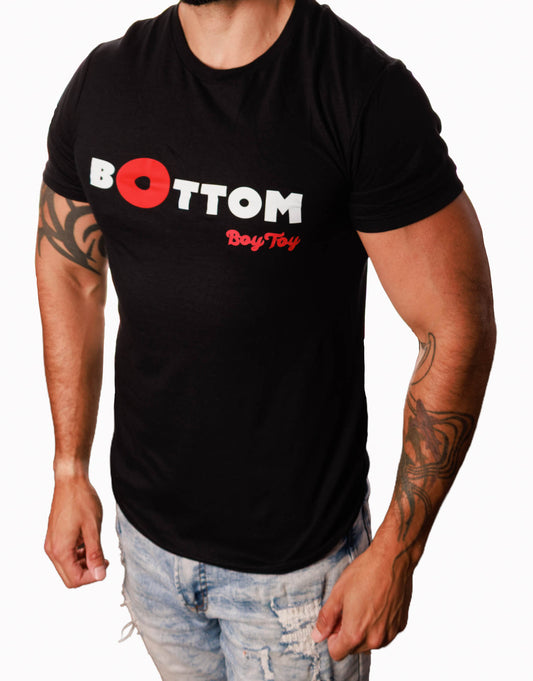 A man wearing a black short-sleeve t-shirt with a graphic on the chest that reads 'BOTTOM BoyToy' in white and red, paired with light-wash distressed jeans against a plain white background.