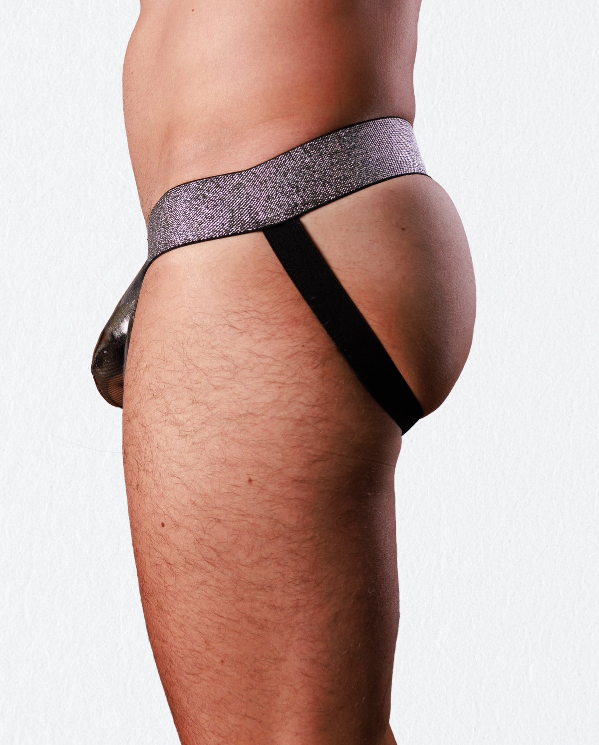Close-up side view of a person wearing a minimal thong-style underwear bottom with a glittery lavender-purple waistband and a black side strap, against a plain light background.