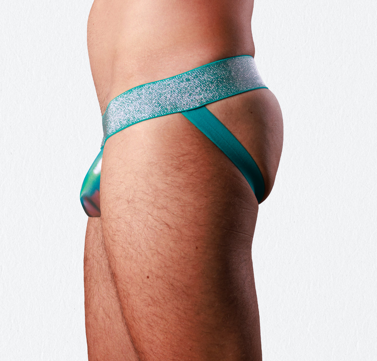 Close-up side view of a person wearing a teal and iridescent jockstrap with a glittery silver waistband, against a plain light background.