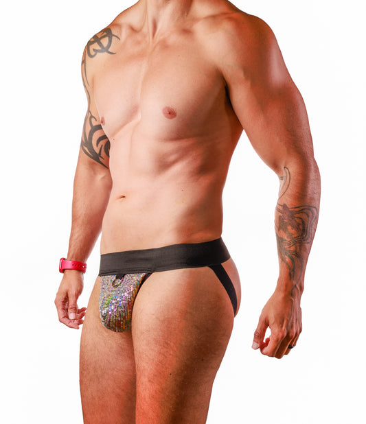 A shirtless male model wearing a low-rise, asymmetrical men's brief. The front panel is covered in a dense, iridescent sequin pattern that shifts colors, while the waistband and side straps are solid black.