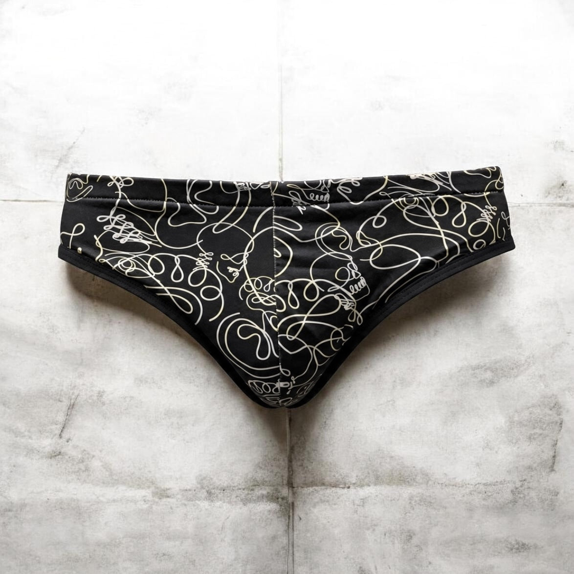 Black bikini-style underwear with an abstract white line pattern, laid flat on a light gray tiled surface.