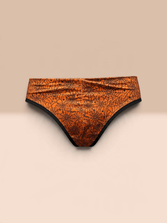 Orange bikini bottoms with a black spiderweb and spider graphic print, displayed on a light wooden surface against a dark gray background.