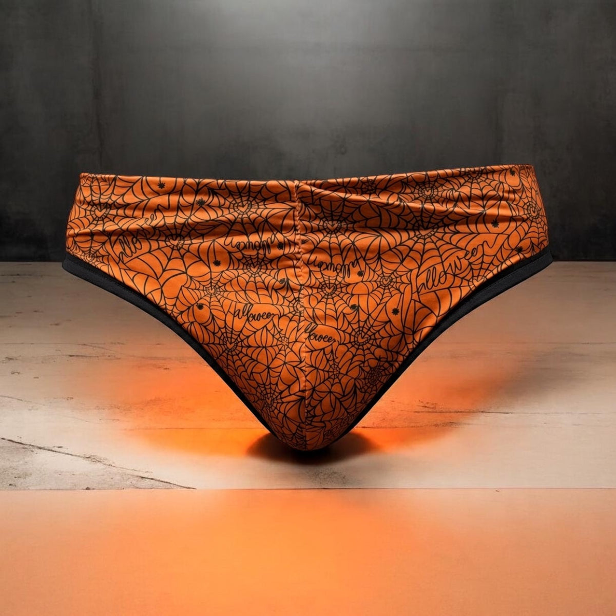 Orange bikini bottoms with a black spiderweb and spider graphic print, displayed on a light wooden surface against a dark gray background.